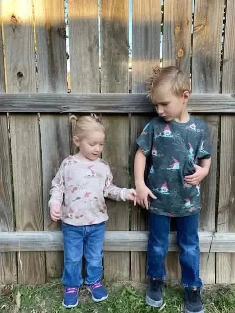Two children wear shirts with a green gnome toile pattern, standing in front of a fence.