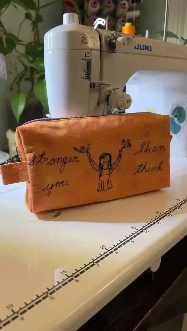 Orange zipper pouch with "Stronger Than You Think" embroidery and girl figure, on sewing table.