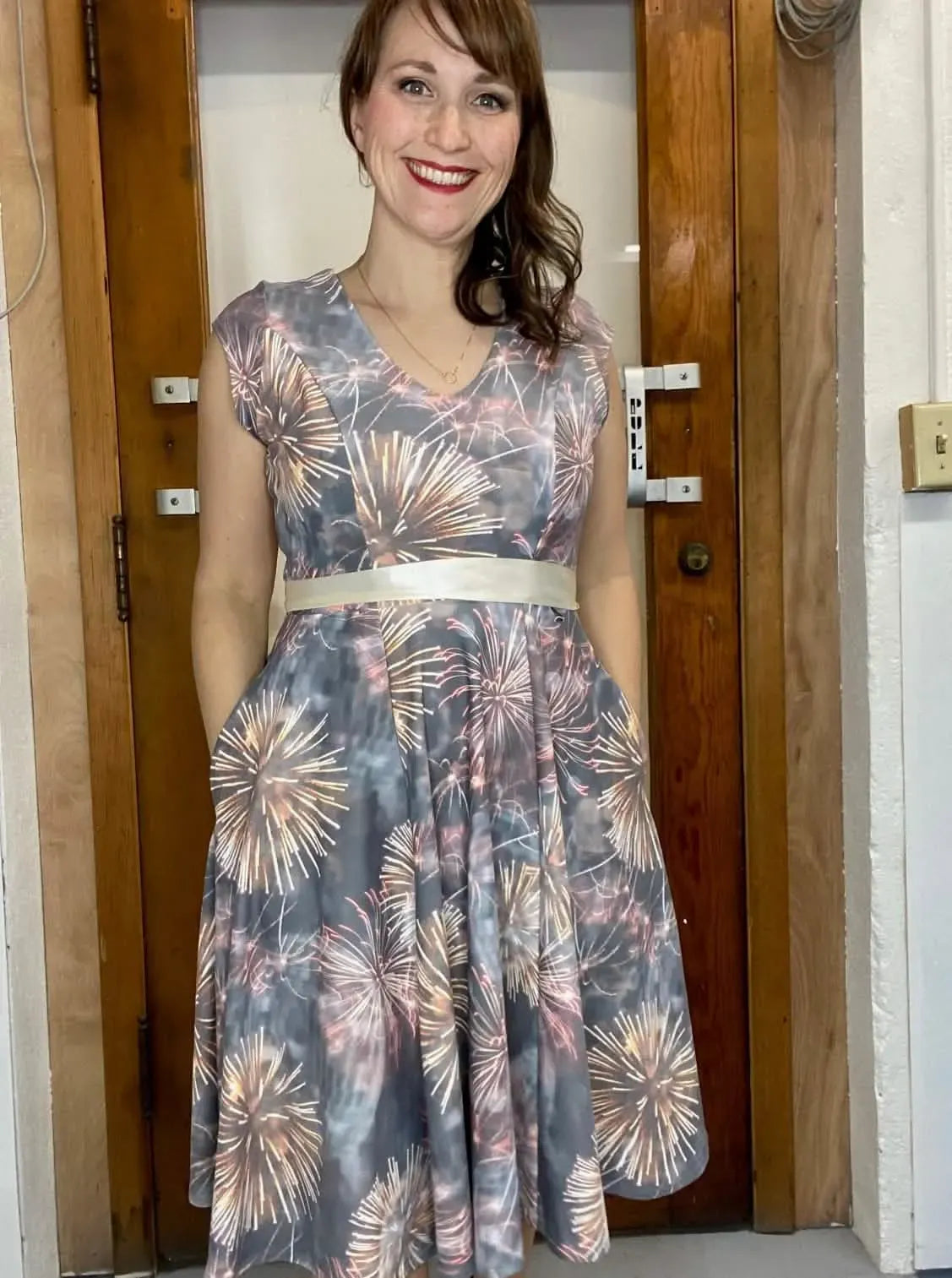 A woman smiles, wearing a grey dress with a firework pattern and a white belt.