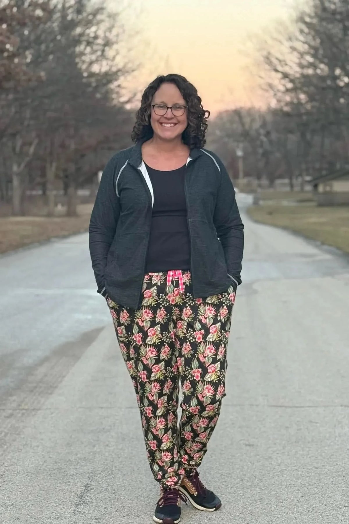 A woman wears black floral pants with pink and green flowers, a black top, and a gray jacket.