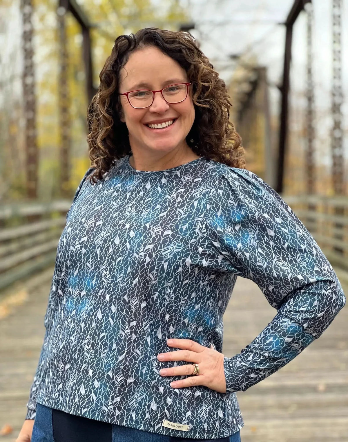 A woman in a blue and white leaf-patterned top smiles on a wooden bridge.