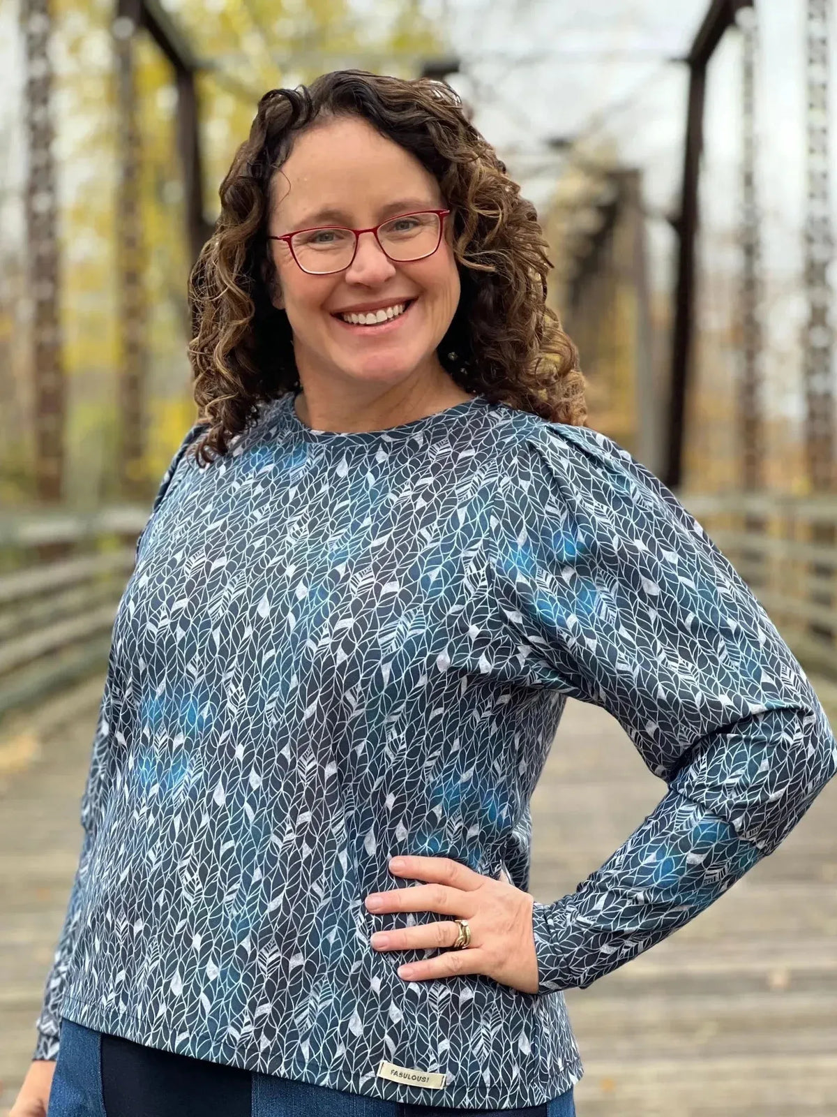 A woman smiles, wearing a long-sleeved top with a silver abstract leaf pattern.