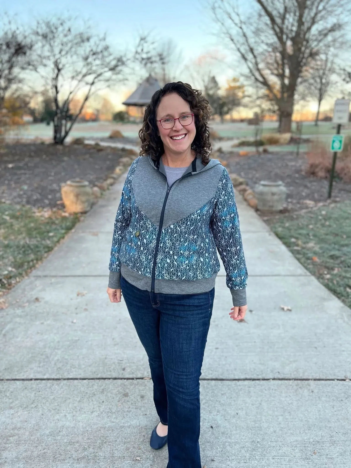 A woman smiles wearing a gray and blue patterned zip-up hoodie and jeans.