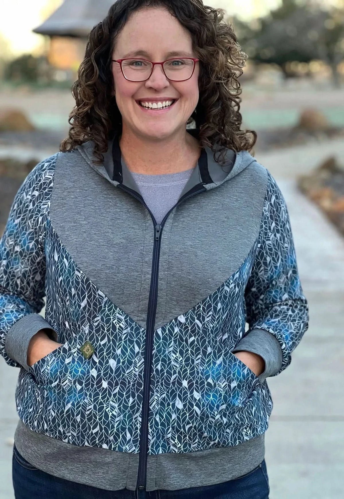 A woman smiles wearing a blue and gray zip-up hoodie with a silver leaf pattern.