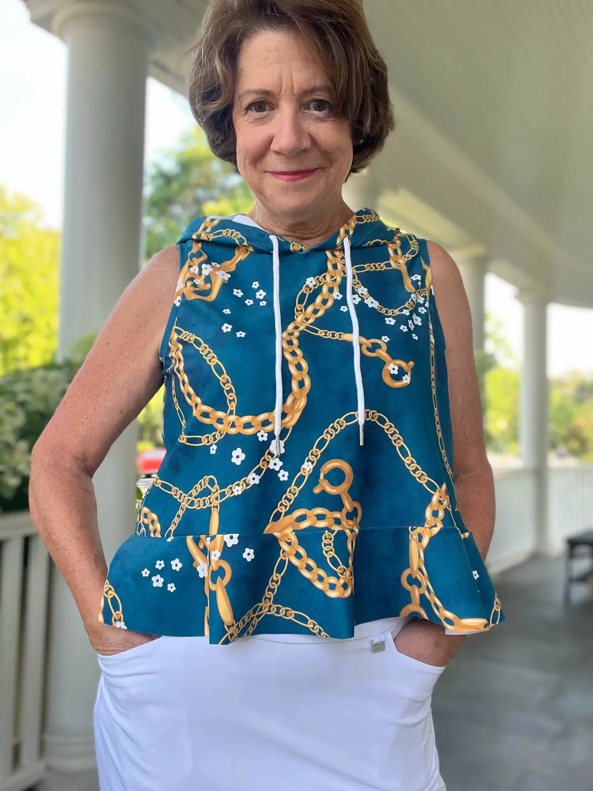 A woman wears a teal top with gold chains and white flowers, and white pants.
