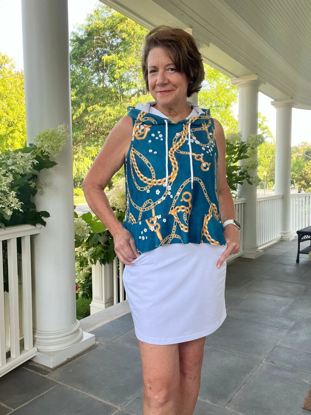 Woman in a teal, chain-patterned sleeveless hoodie and white skirt.