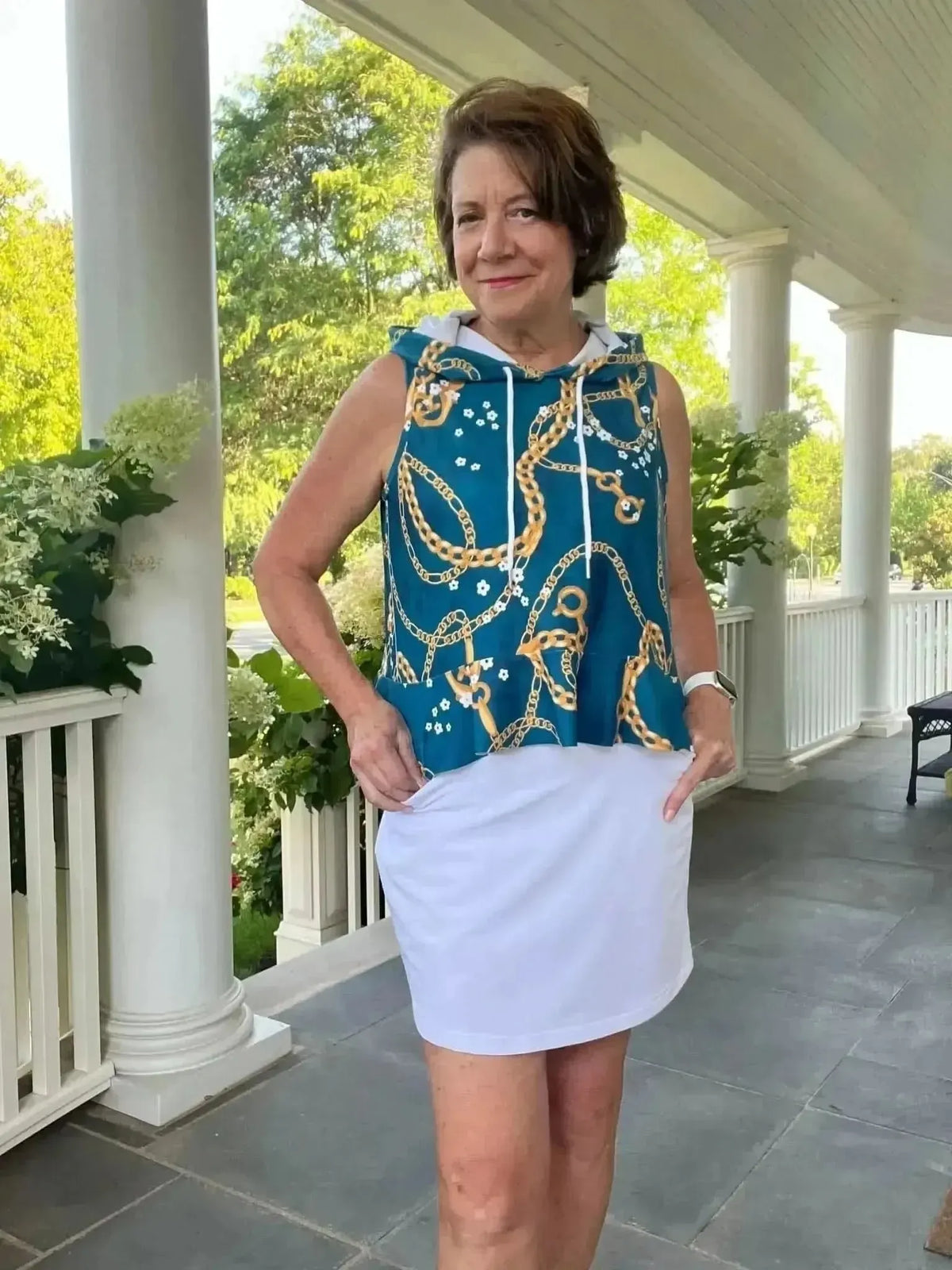 Woman modeling a sleeveless teal hoodie with floral chain print over a white skort on a porch.