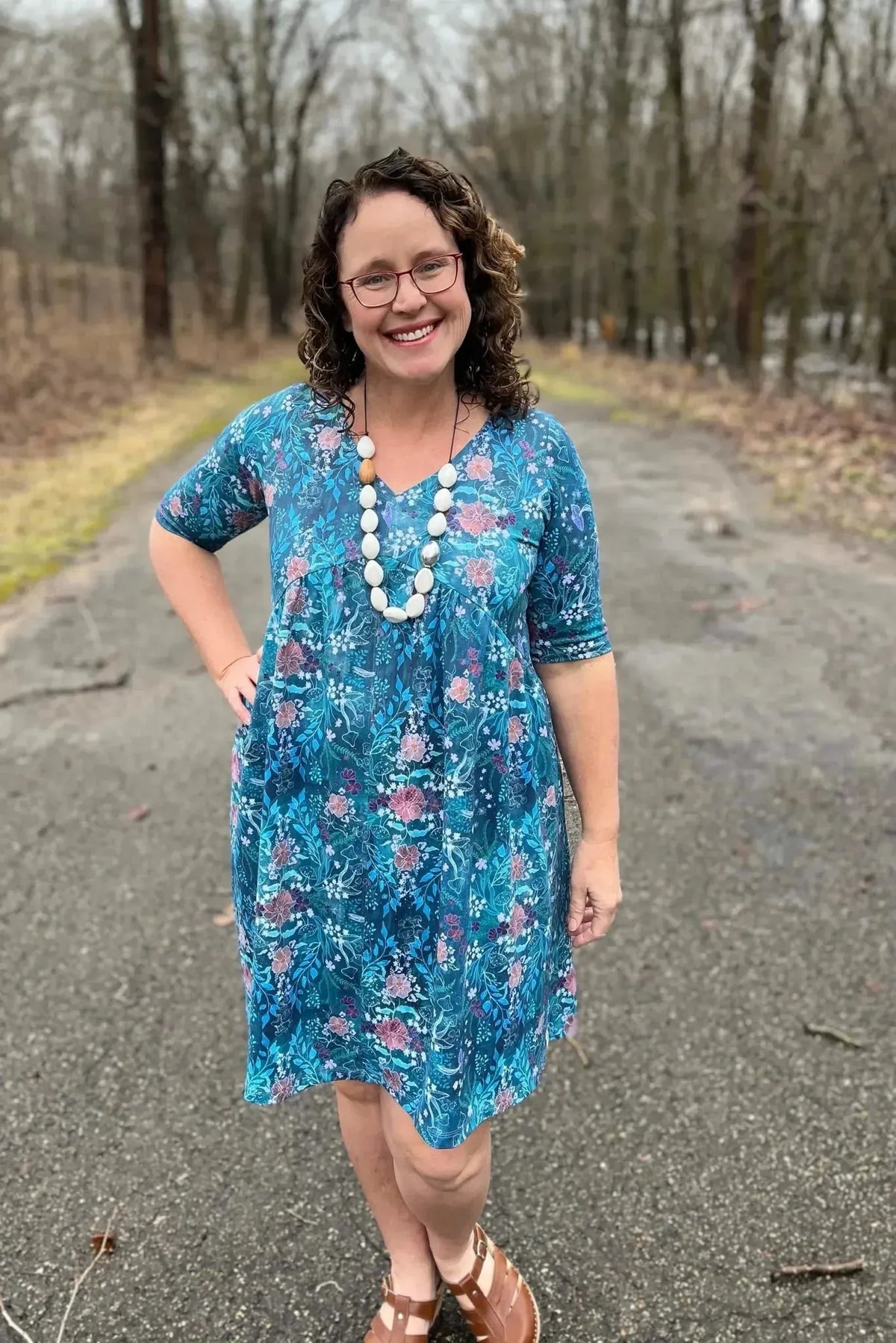 Woman modeling a teal woodland print dress made from Enchanted Glen fabric on a wooded path.