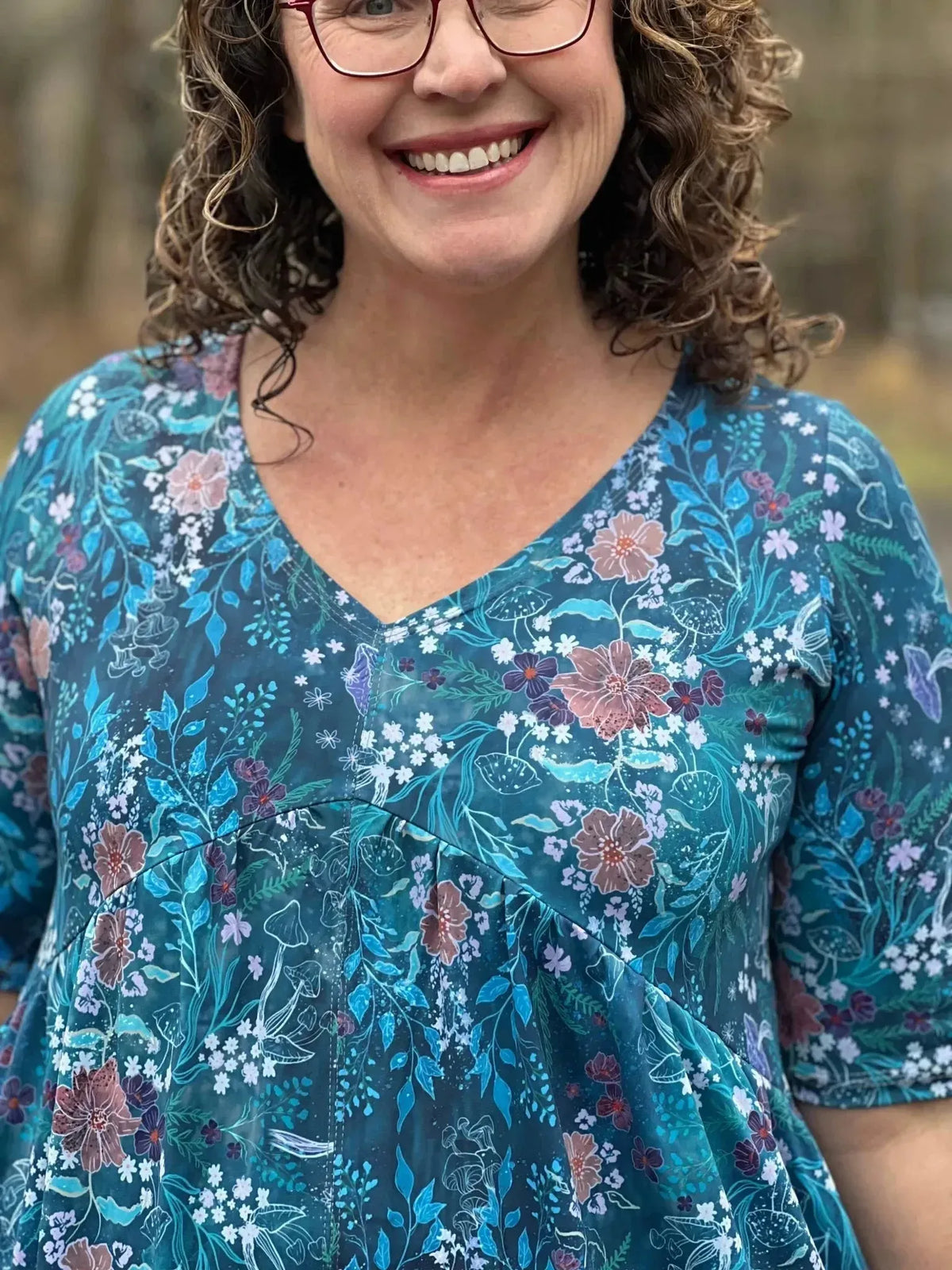 A woman smiles, wearing a teal floral print top with a v-neck.