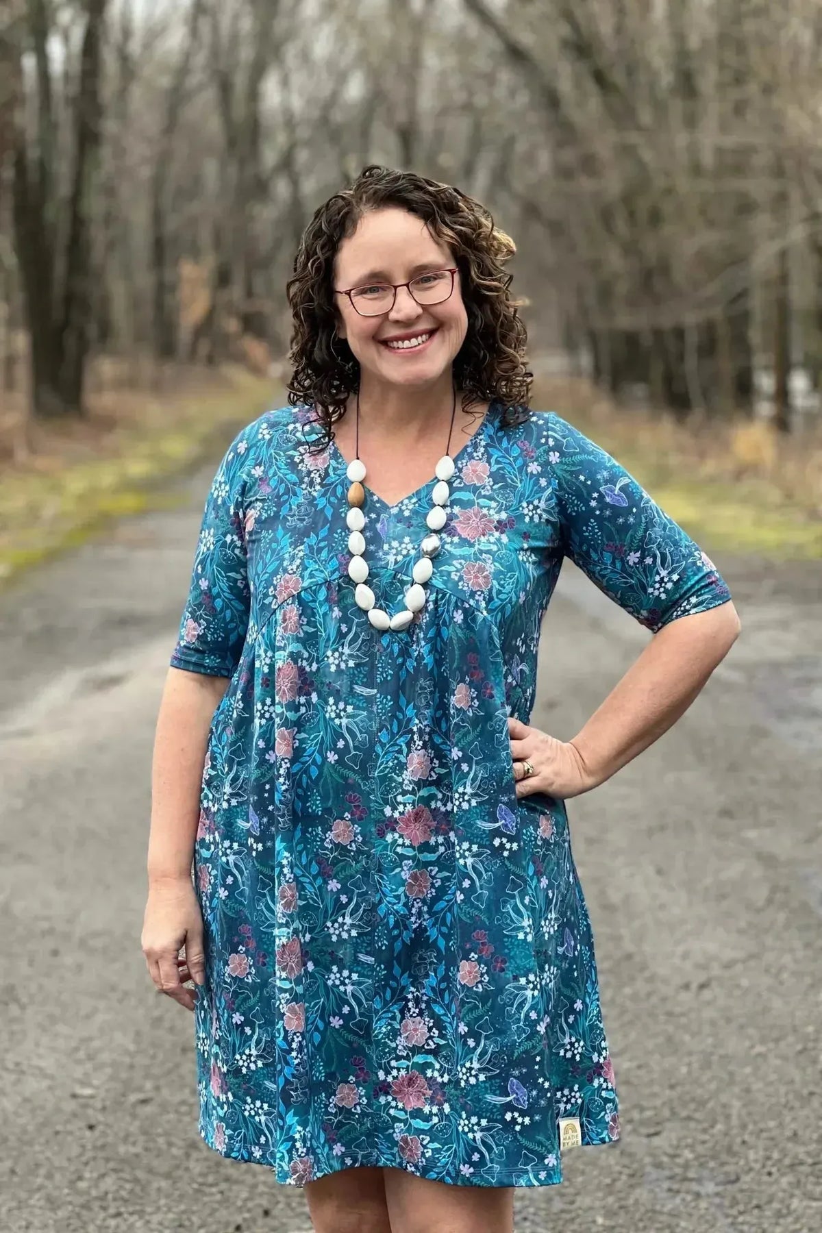 Woman modeling a teal woodland print dress with floral patterns on a wooded path.