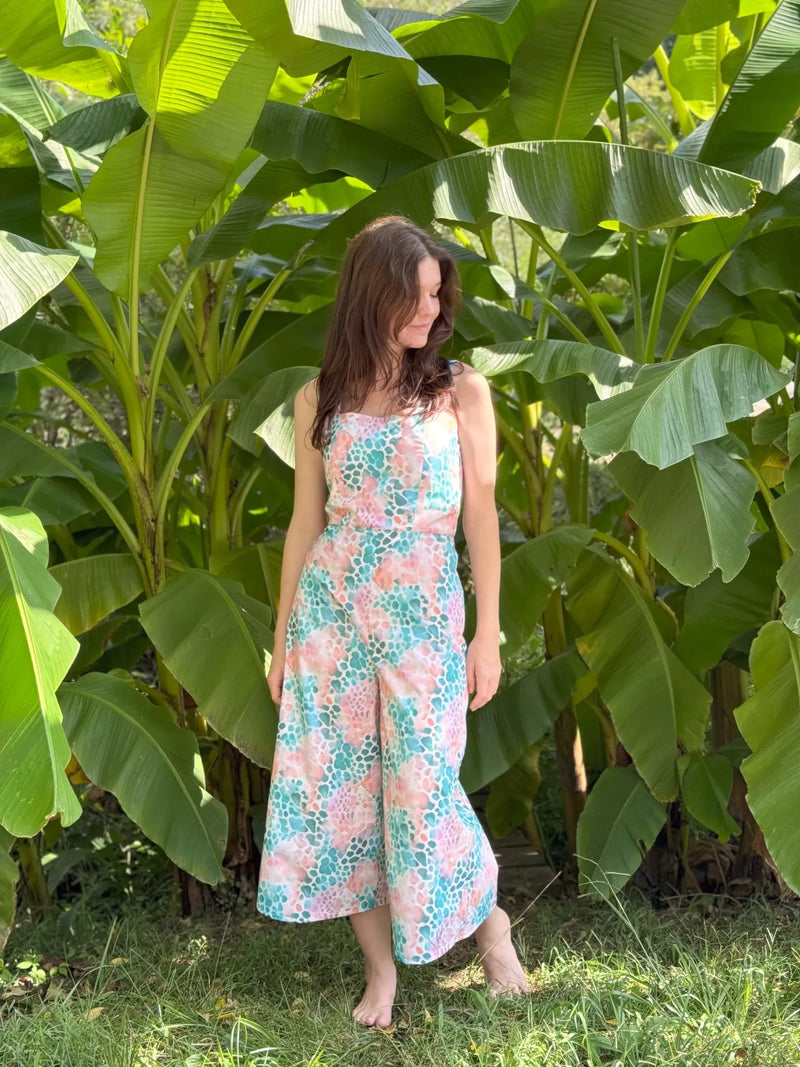 A woman in a colorful, patterned jumpsuit stands barefoot in front of large green leaves.