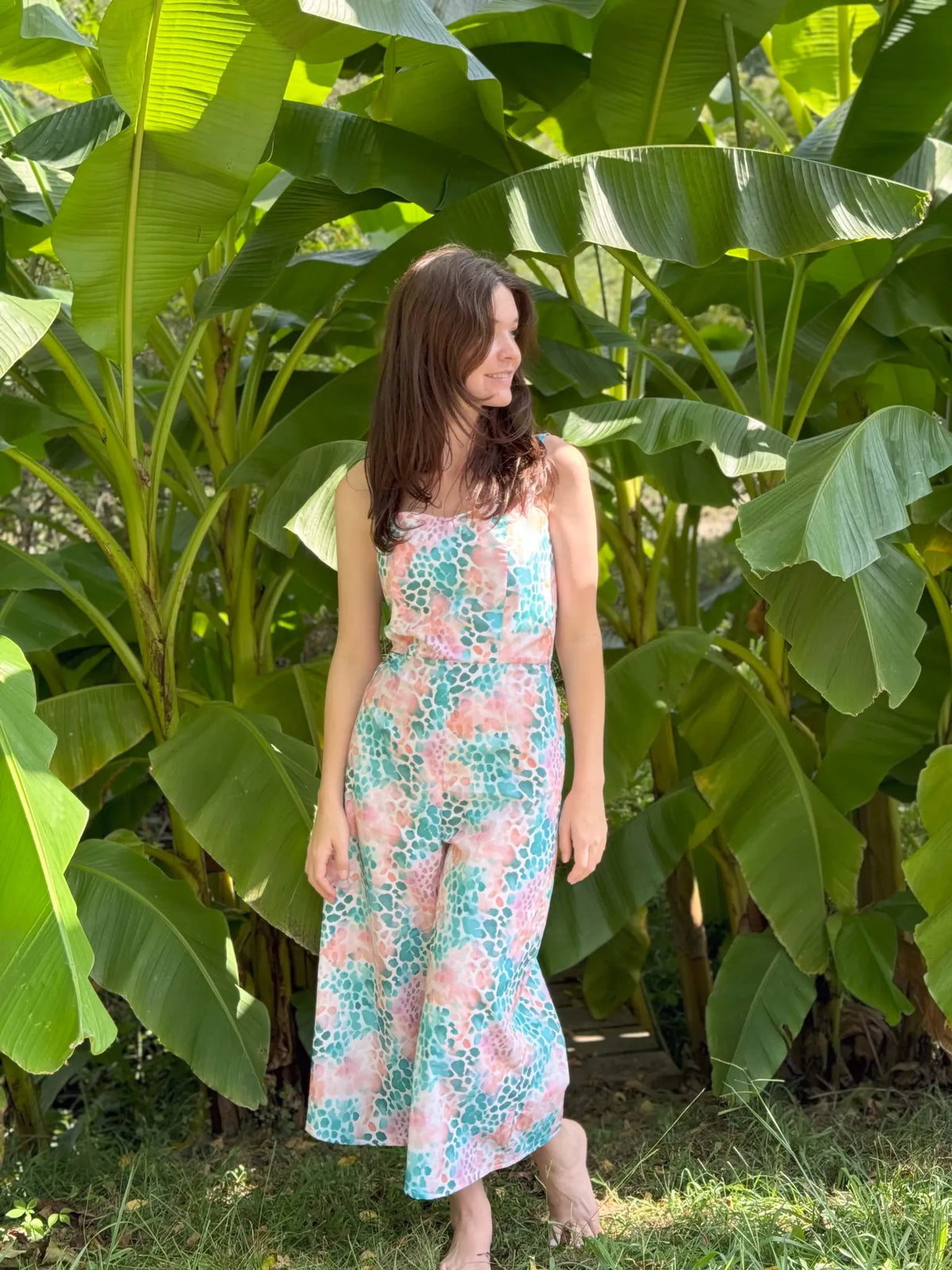 A woman in a colorful, patterned jumpsuit stands barefoot among large green leaves.