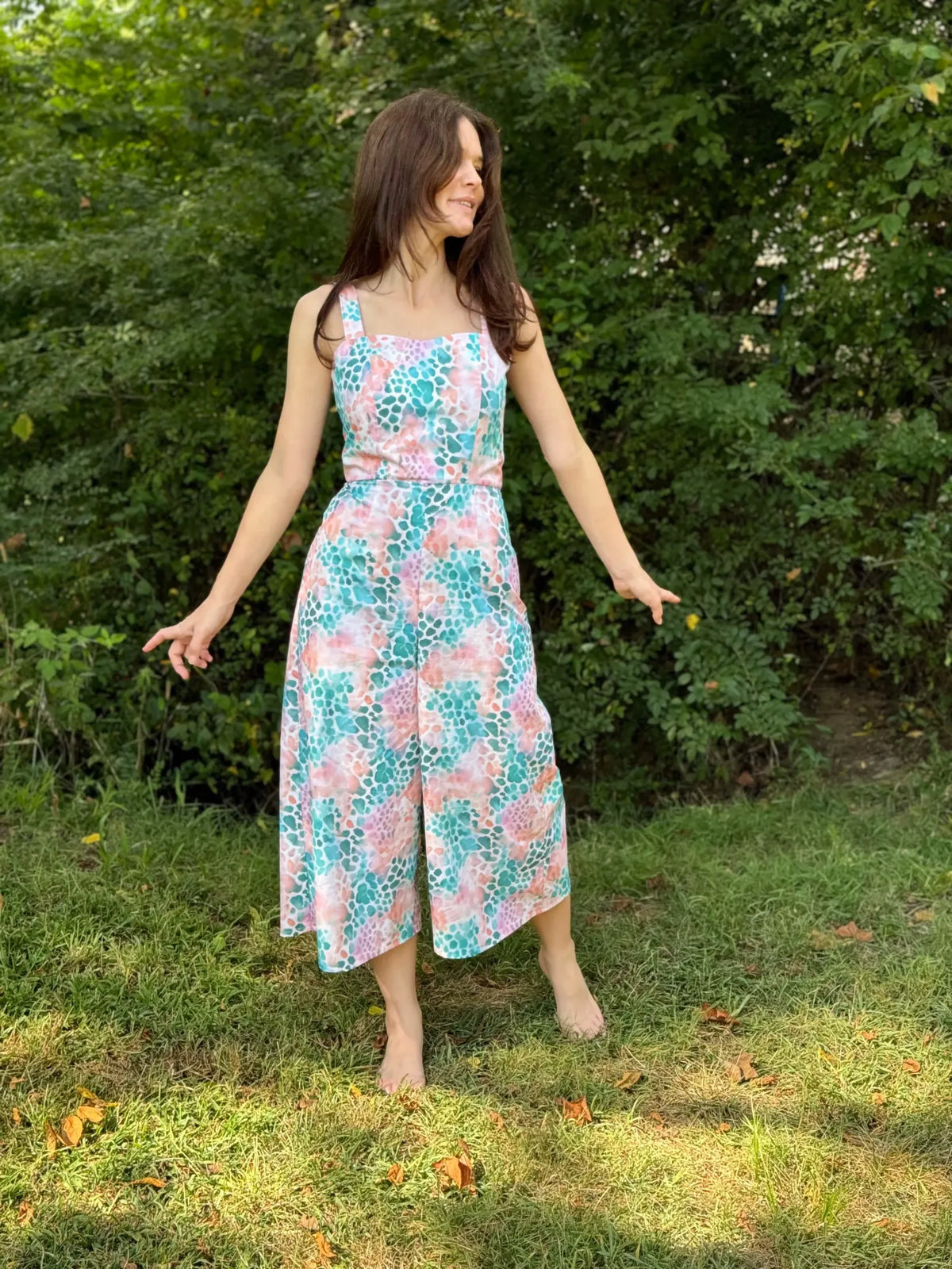 A woman in a colorful, patterned jumpsuit stands barefoot in grass.