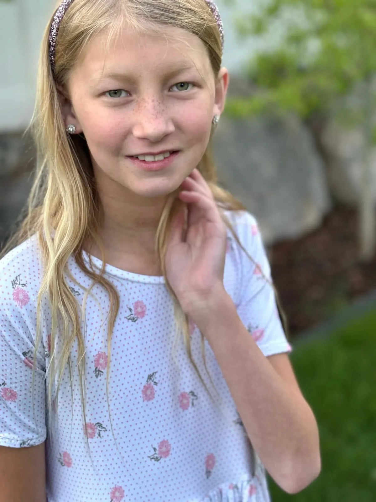 A young girl with blonde hair and a sparkly headband wears a white shirt with pink floral print.