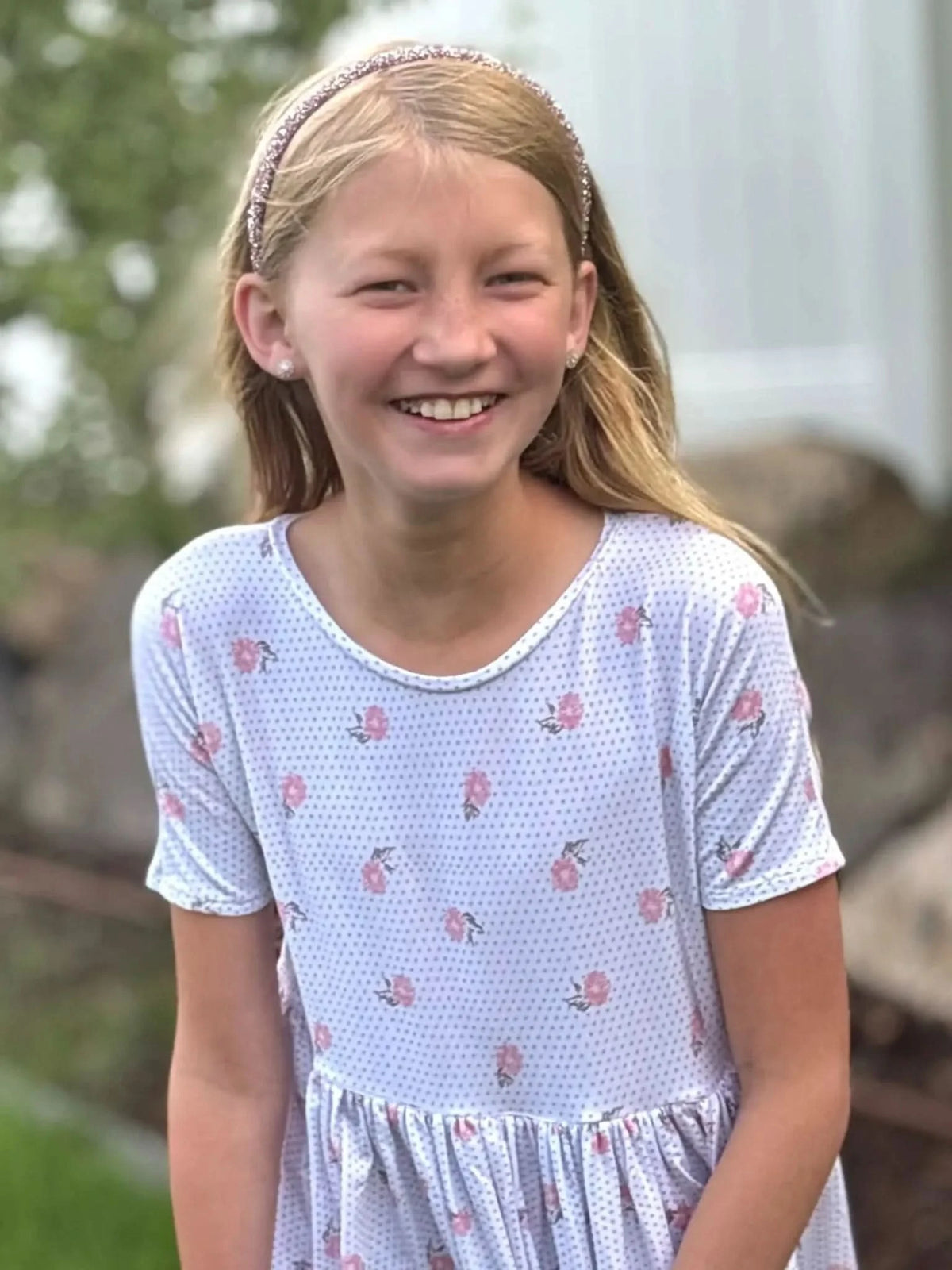 A young girl smiles, wearing a white dress with pink floral and polka dot patterns.