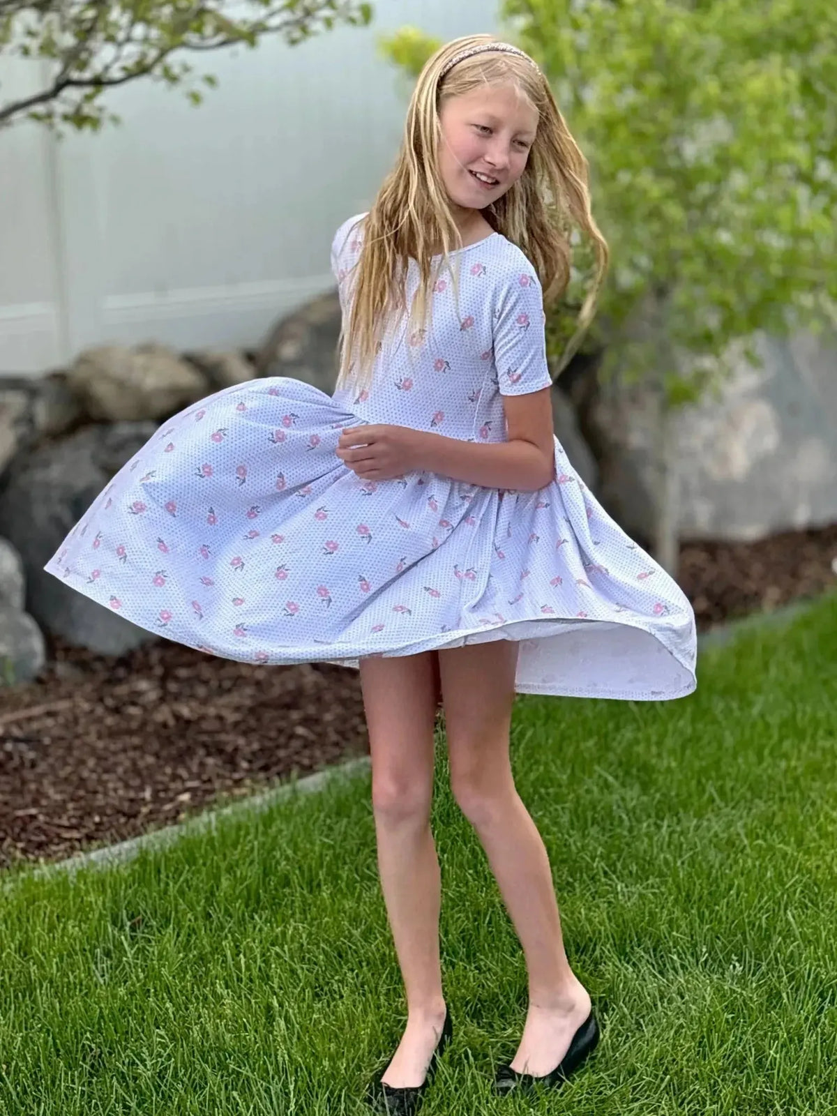 Girl twirling in a white dress with pink vintage floral print, outdoors on grass.