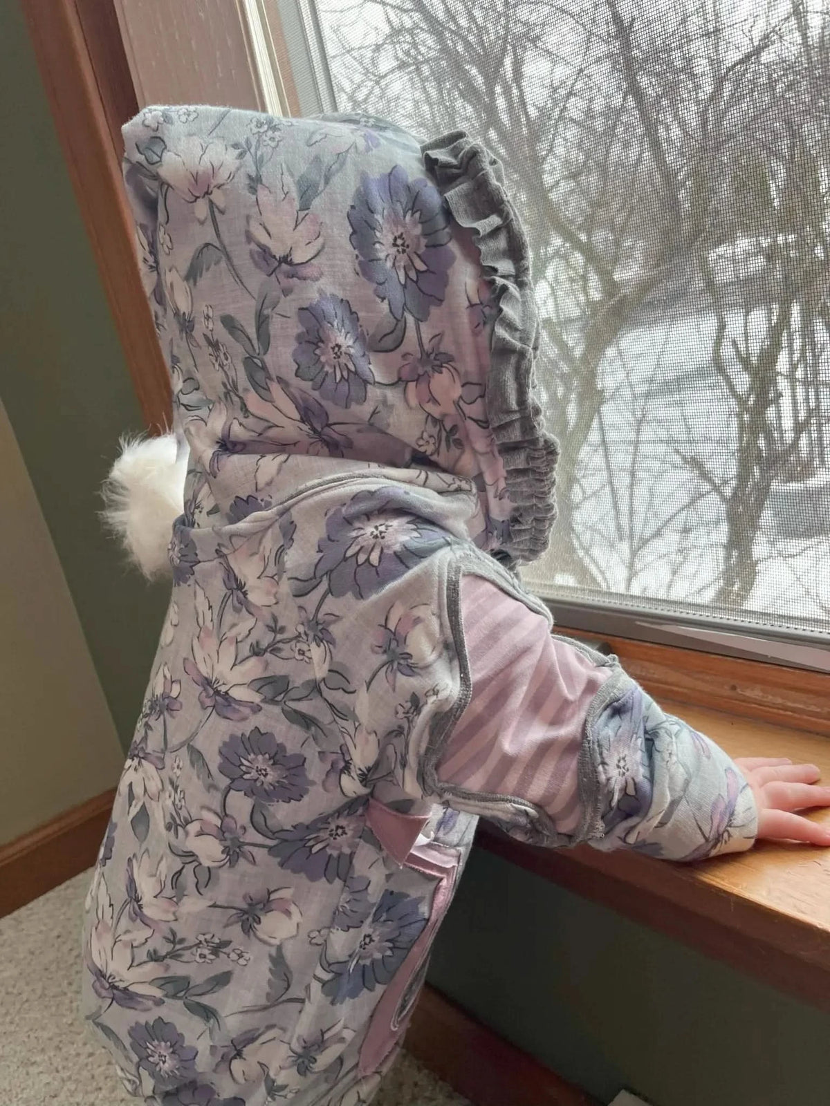 Child in a floral print jacket with a fluffy white accent looks out a window.