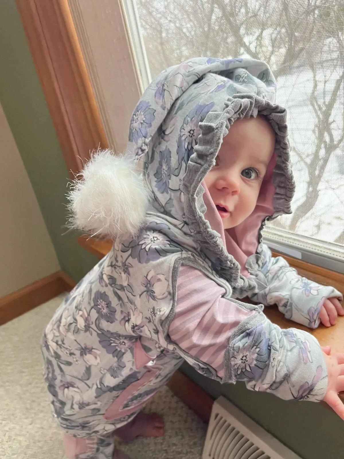A baby in a purple floral and pink striped hooded outfit with a white pompom.