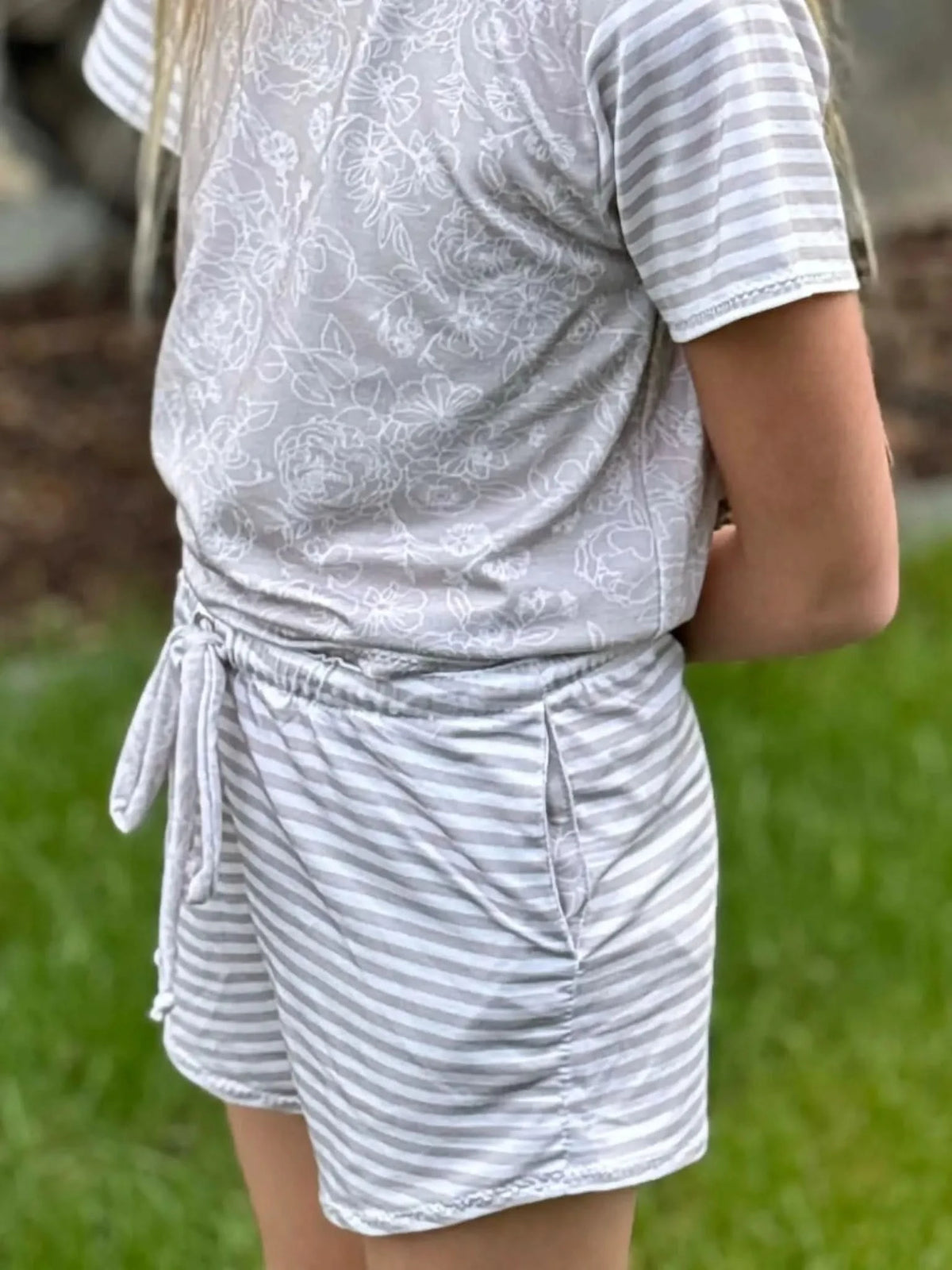 A person wears a light gray romper with a floral top and striped shorts.