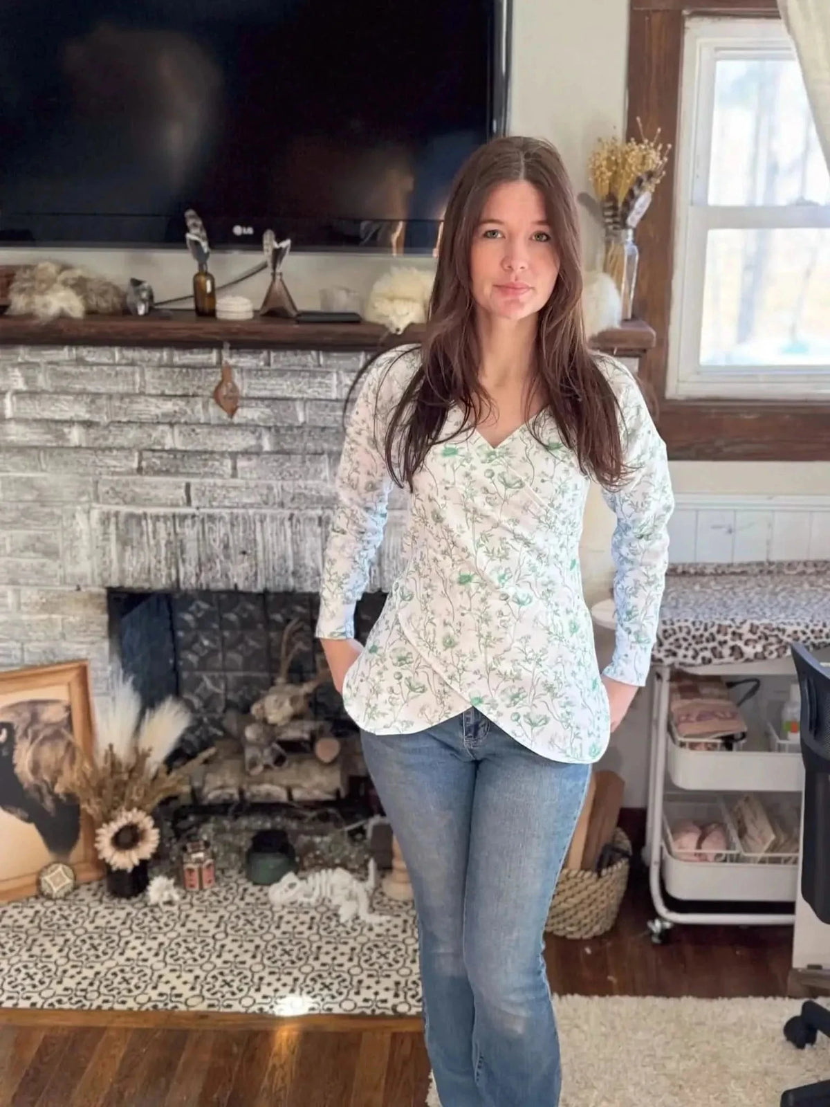 Woman wearing a white wrap blouse with green floral print, styled with jeans in a cozy living room.