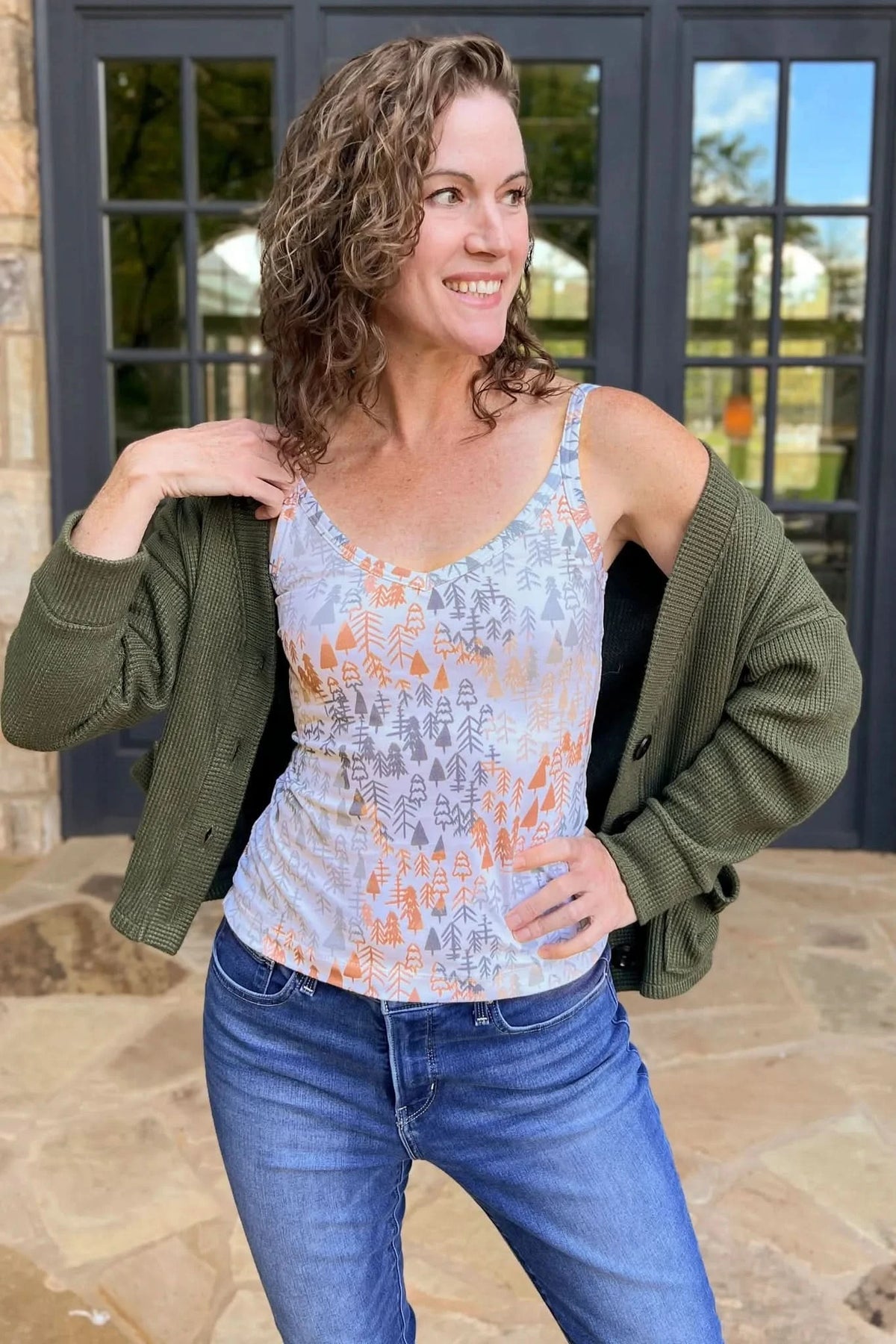 A woman wears a tank top with a light fall trees pattern and a green cardigan.