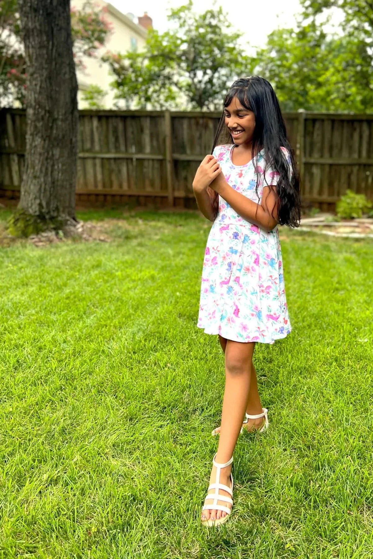 A young girl in a floral dress and white sandals stands in a grassy backyard.