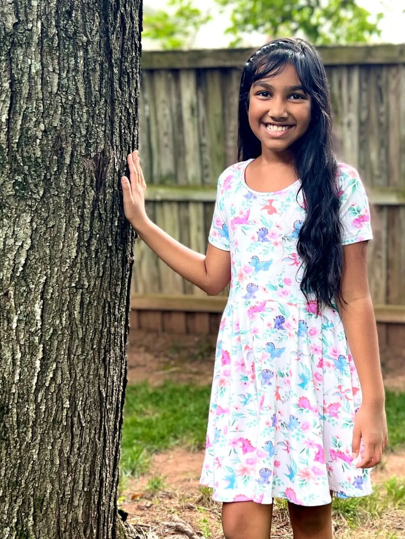 A young girl in a white dress with pink and blue dinosaur and floral patterns.