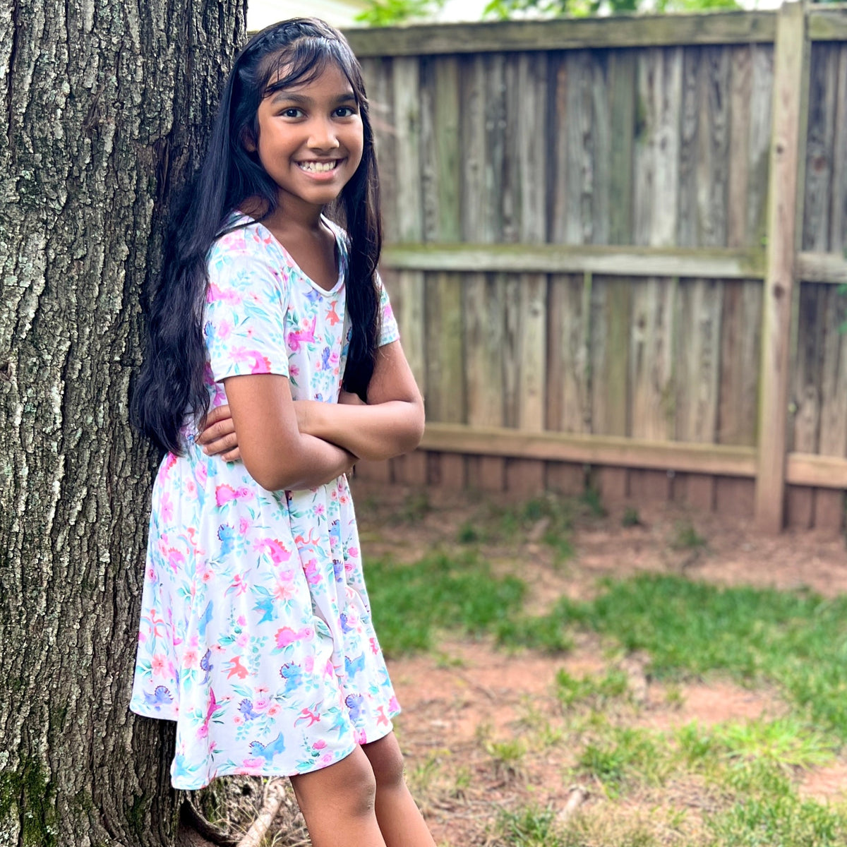 Young girl in a floral dress with dinosaurs standing next to a tree in a backyard.