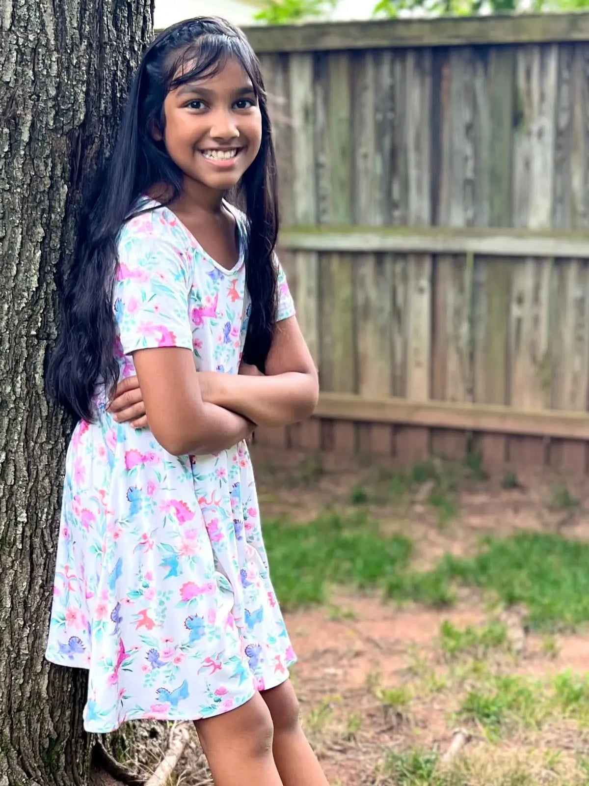 A young girl in a floral and dinosaur print dress leans against a tree.