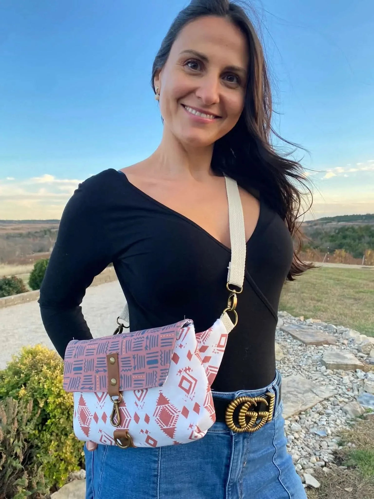 A woman smiles, wearing a black top, jeans, and a crossbody bag with a geometric pattern.