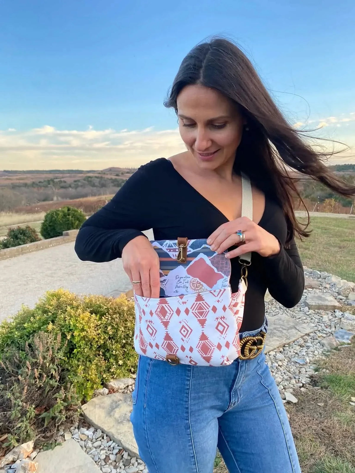 A woman in a black top and jeans holds a Desert Flame patterned bag outdoors.