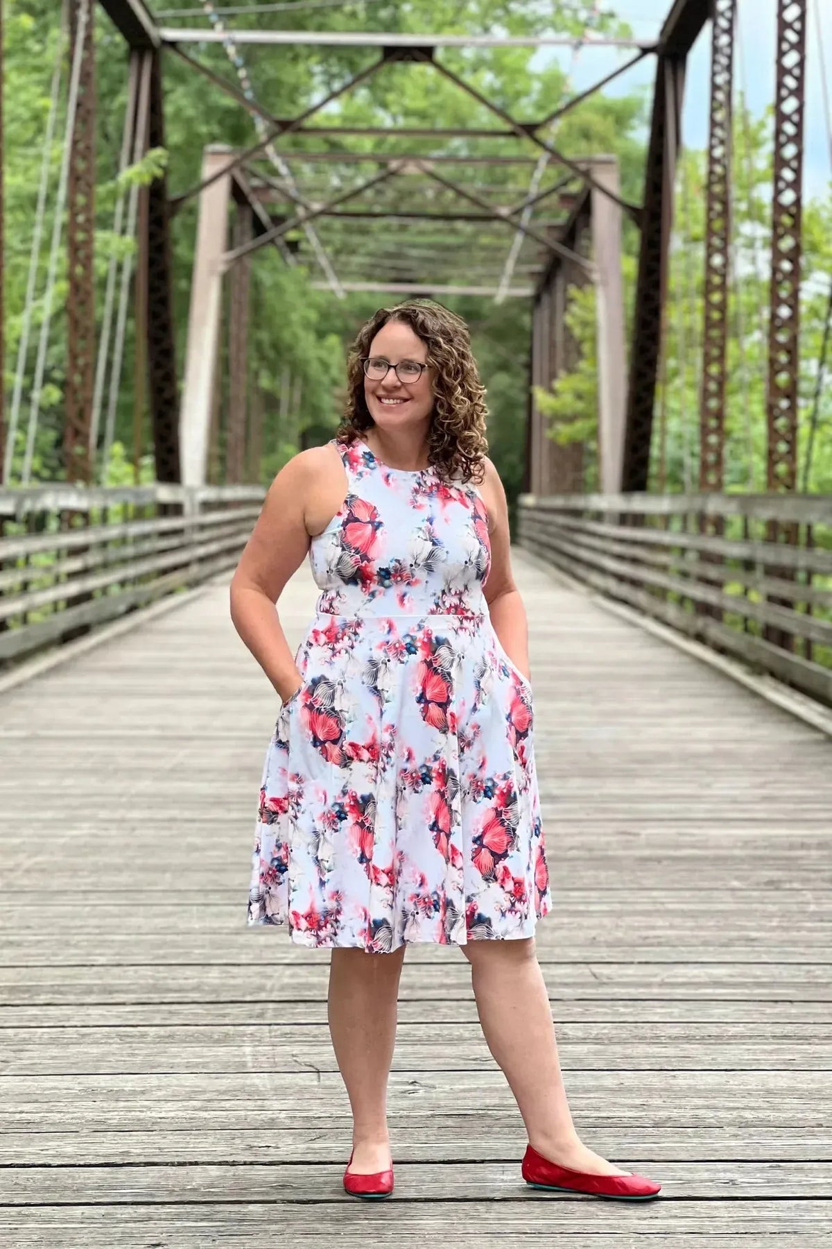 A woman in a floral dress with red and blue flowers stands on a wooden bridge.