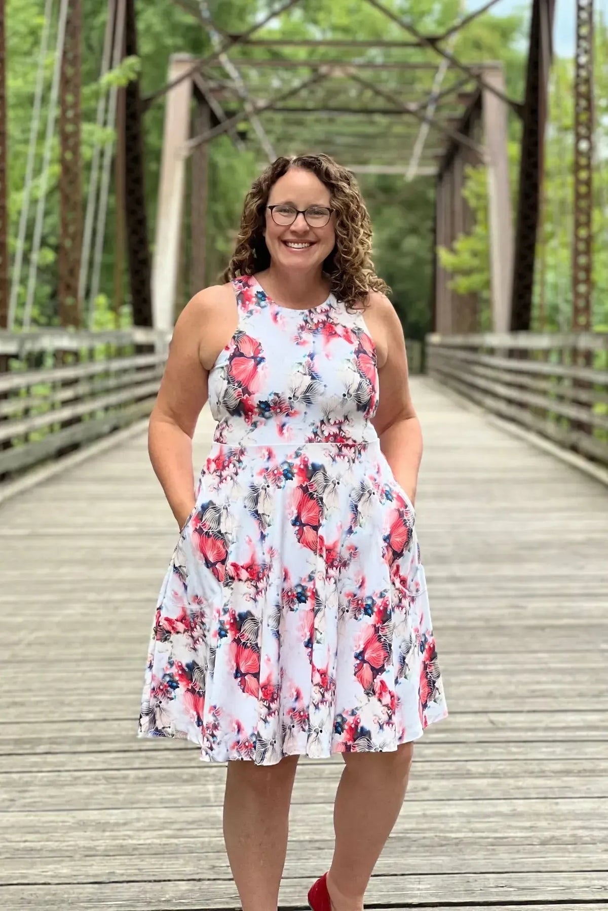 A woman in a floral dress with red and white flowers stands on a wooden bridge.