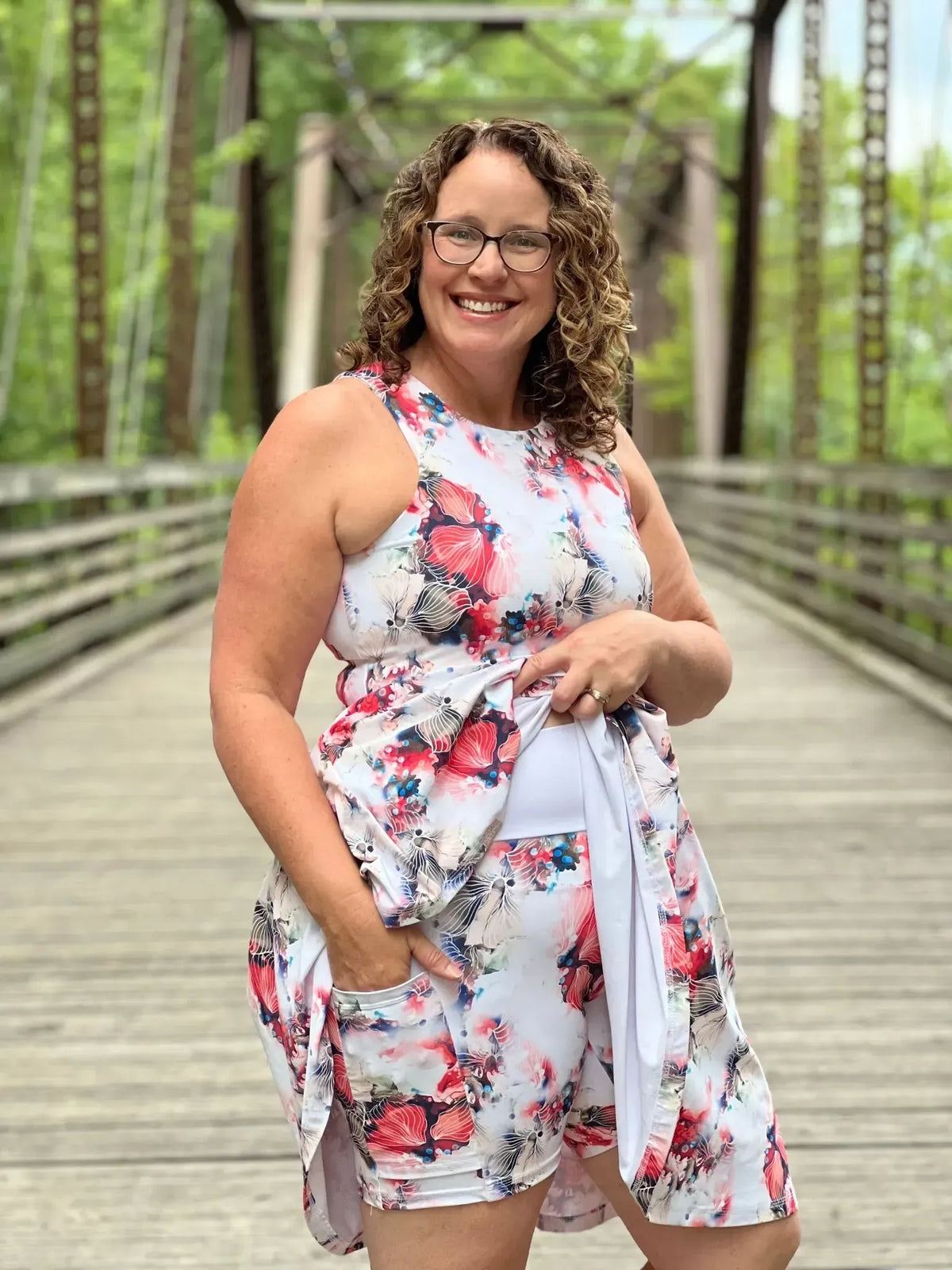 A woman in a floral print dress with pockets stands on a wooden bridge.