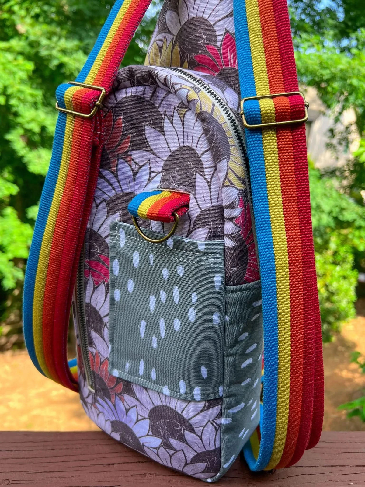 A sunflower-patterned bag with a polka dot pocket and rainbow straps rests on a wooden surface.
