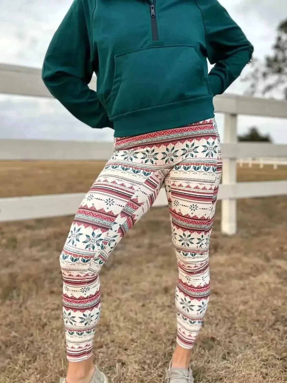 Woman modeling green hoodie and fair isle snowflake leggings outdoors by a white fence.