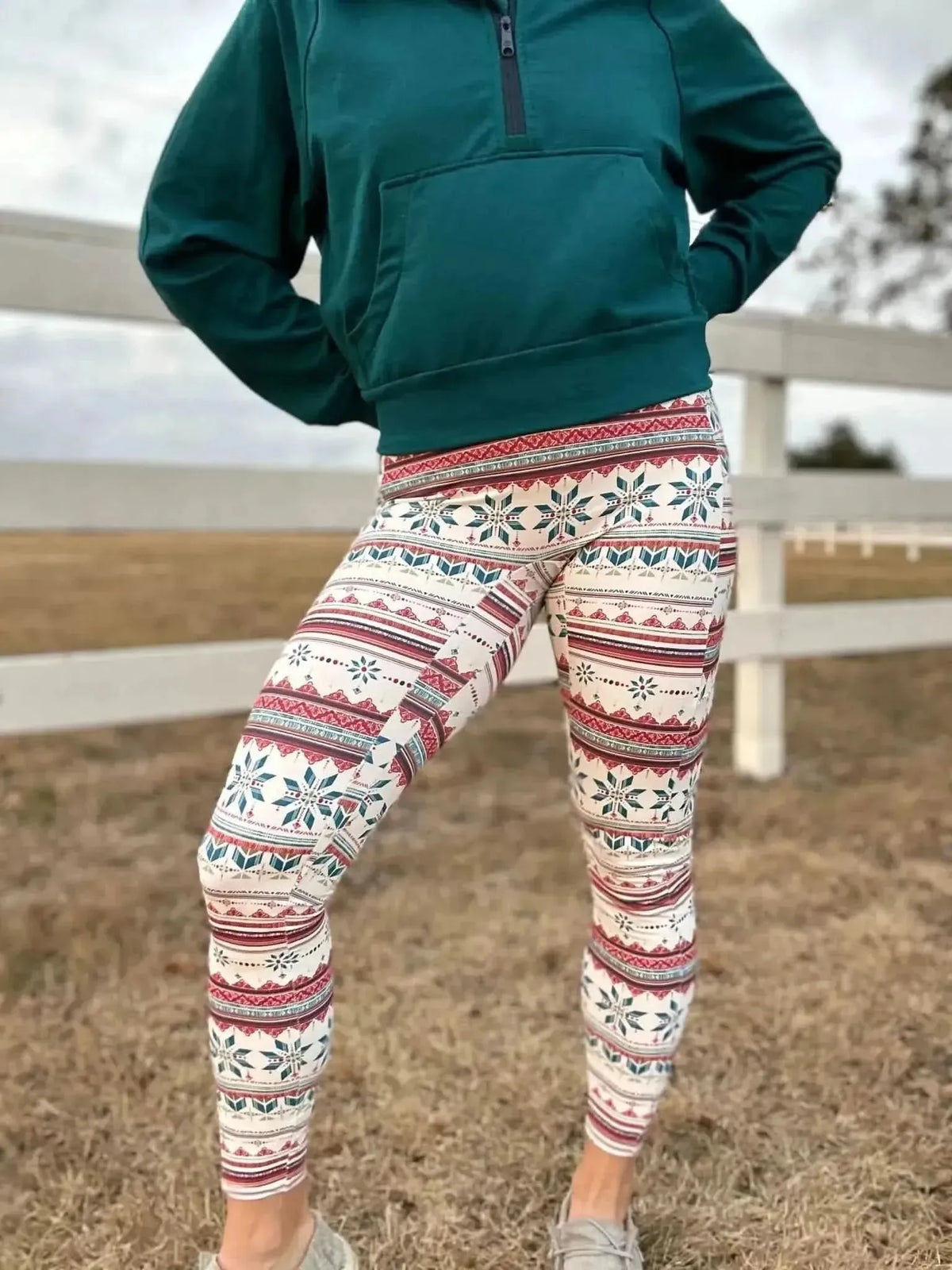 Woman modeling green hoodie and fair isle snowflake leggings outdoors by a white fence.