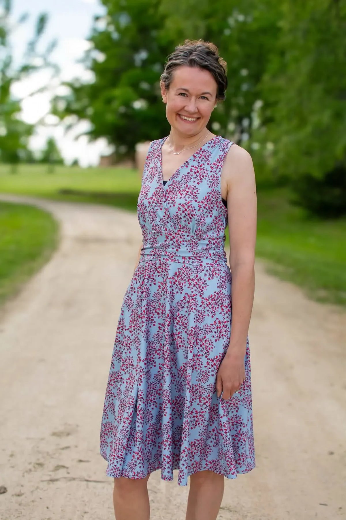 A woman in a light blue dress with a red floral pattern smiles on a dirt path.