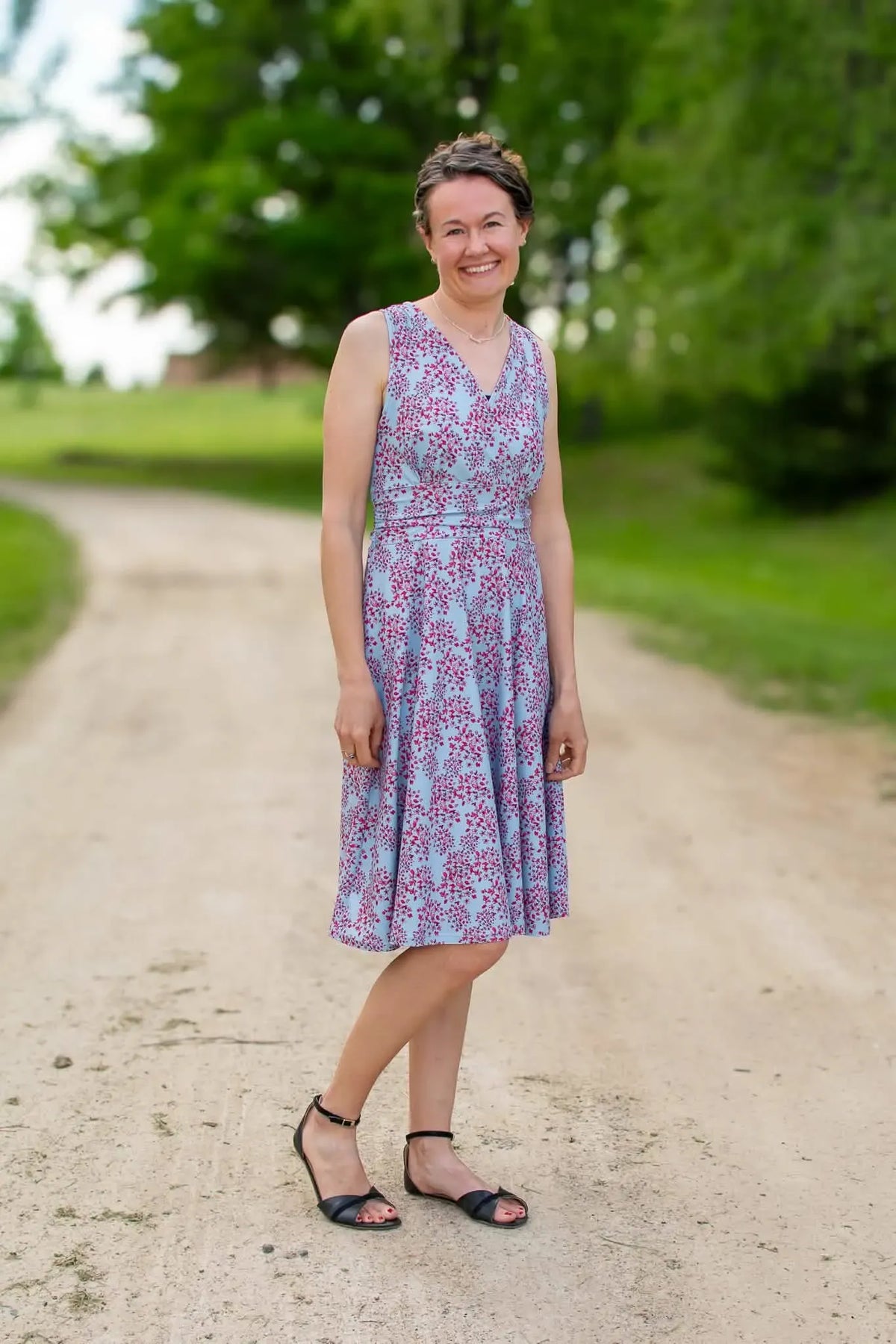 A woman in a light blue dress with red floral patterns stands on a dirt path.
