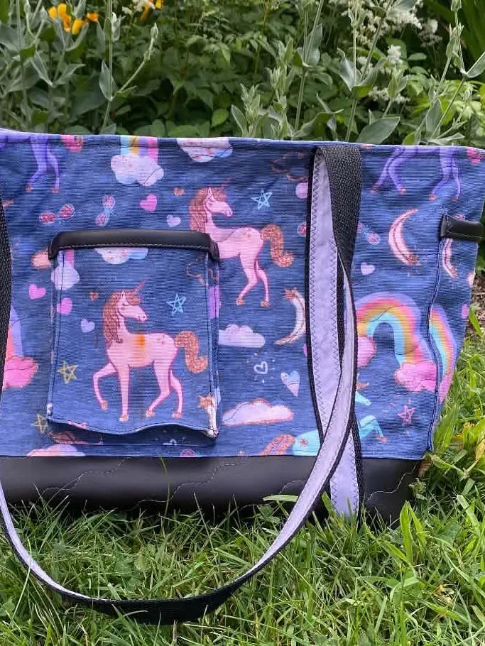 A navy unicorn tote bag with a front pocket sits on green grass.