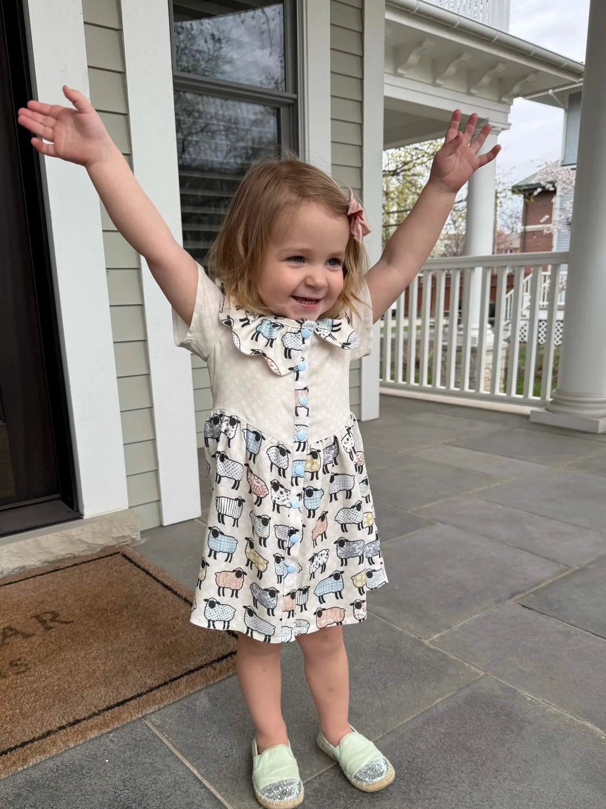 A young girl in a dress with a patchwork sheep pattern stands on a porch.