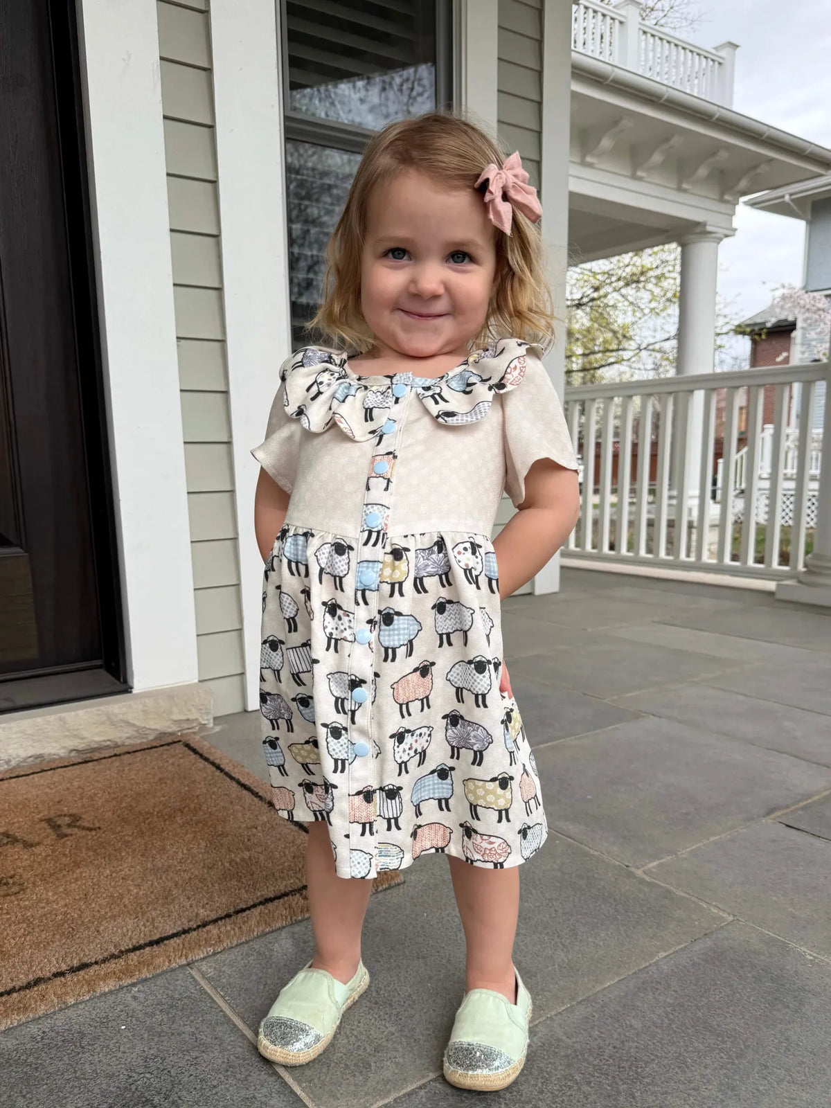 A young girl in a dress with a patchwork sheep pattern stands on a porch.