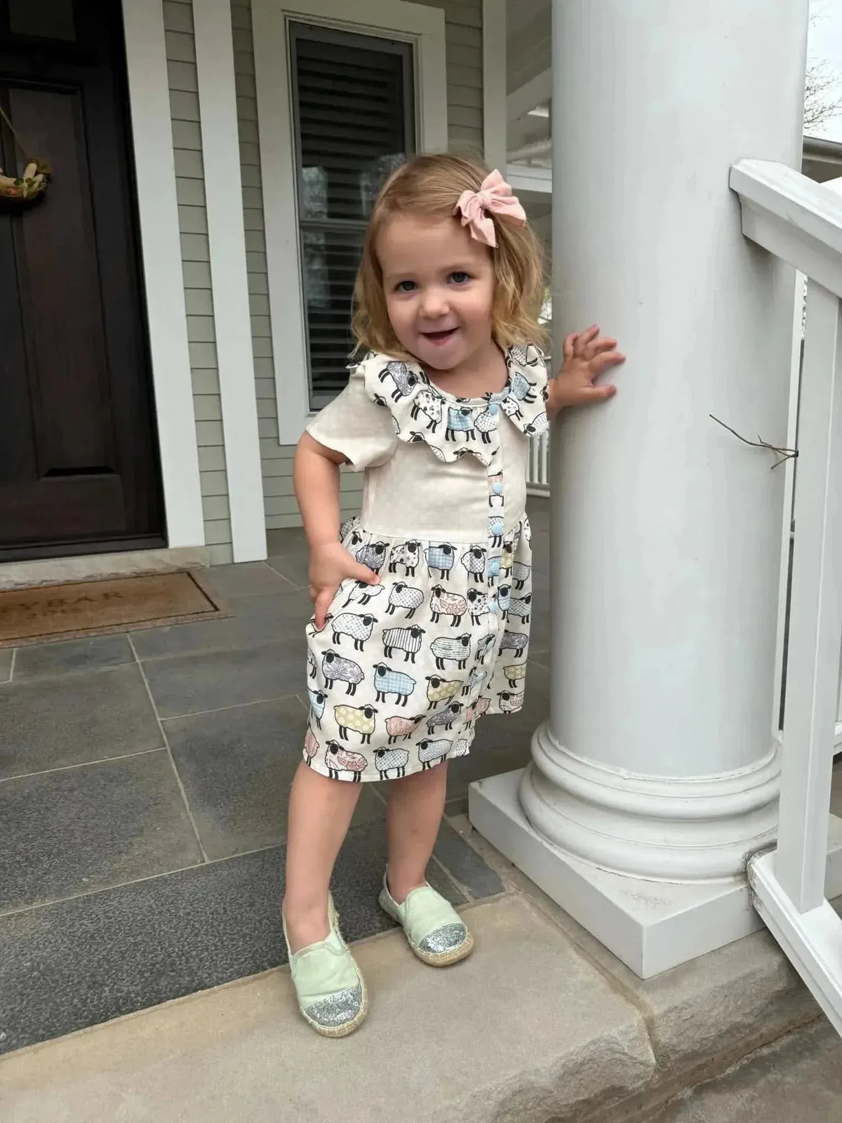 Toddler girl in a short patchwork sheep print dress on a porch.