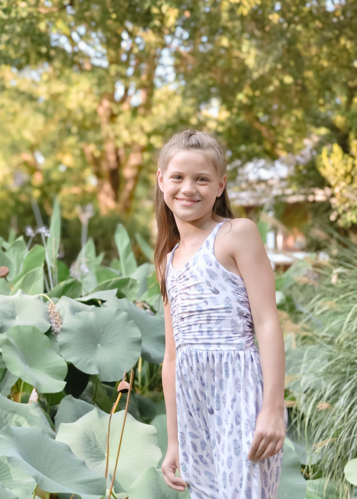 A young girl with blonde hair stands in a garden pond wearing a white sleeveless halter dress with a light blue abstract pattern. Large green lotus leaves fill the foreground, with a tree and greenery in the background.