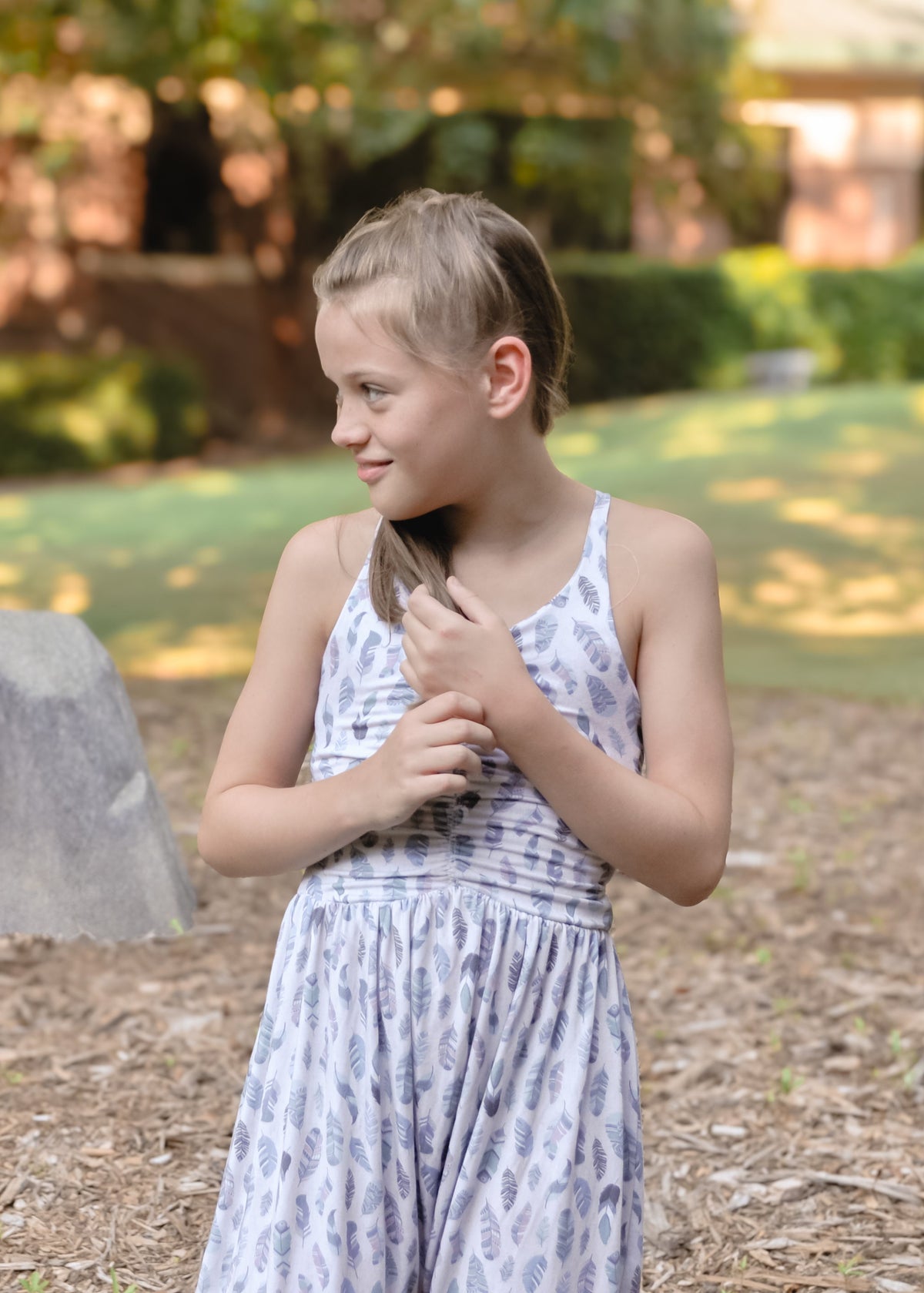 A young girl with blonde hair tied back stands outdoors in a grassy yard wearing a sleeveless white dress with a light blue abstract pattern. She is looking to the side while holding a small black object near her chest.