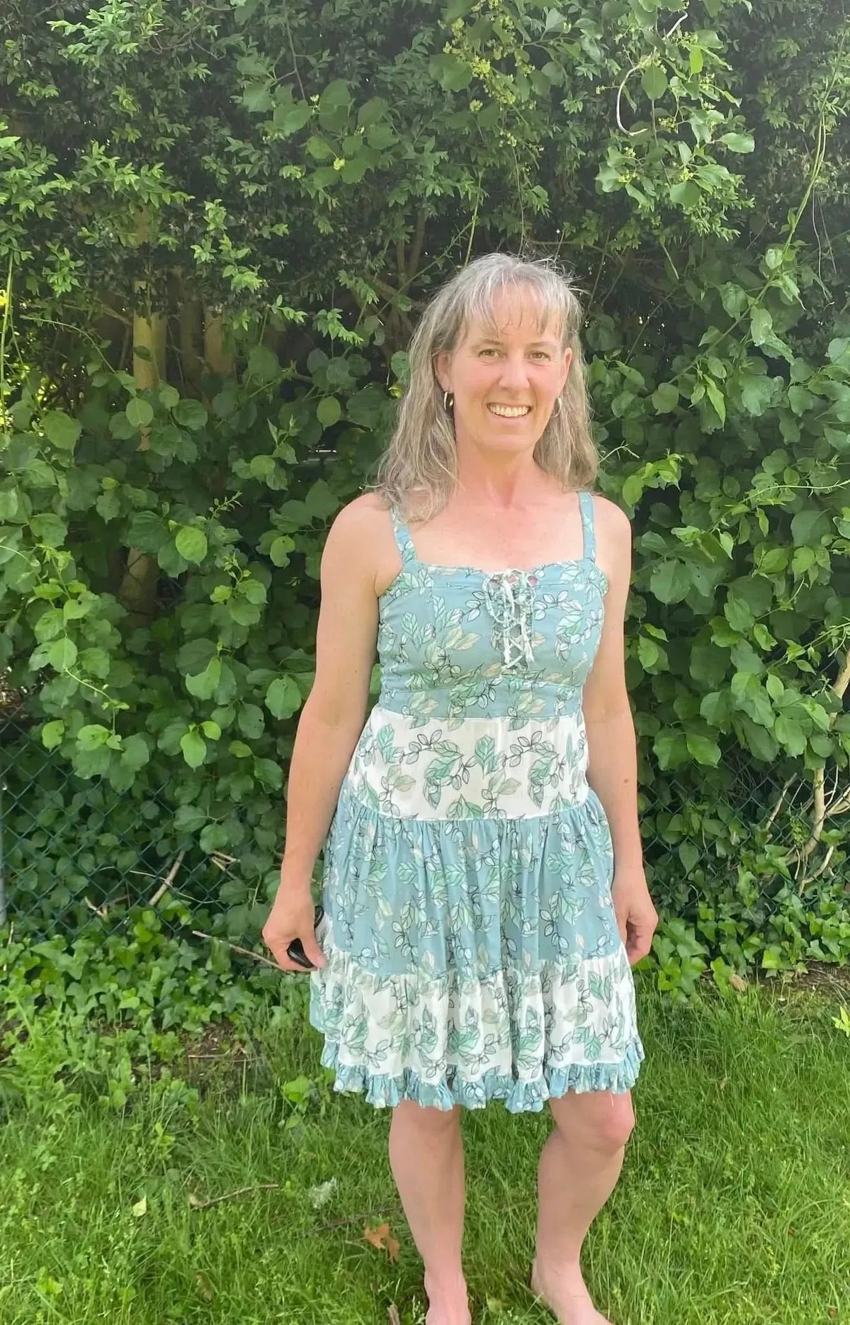 A woman in a light blue and white floral dress stands barefoot in front of green foliage.