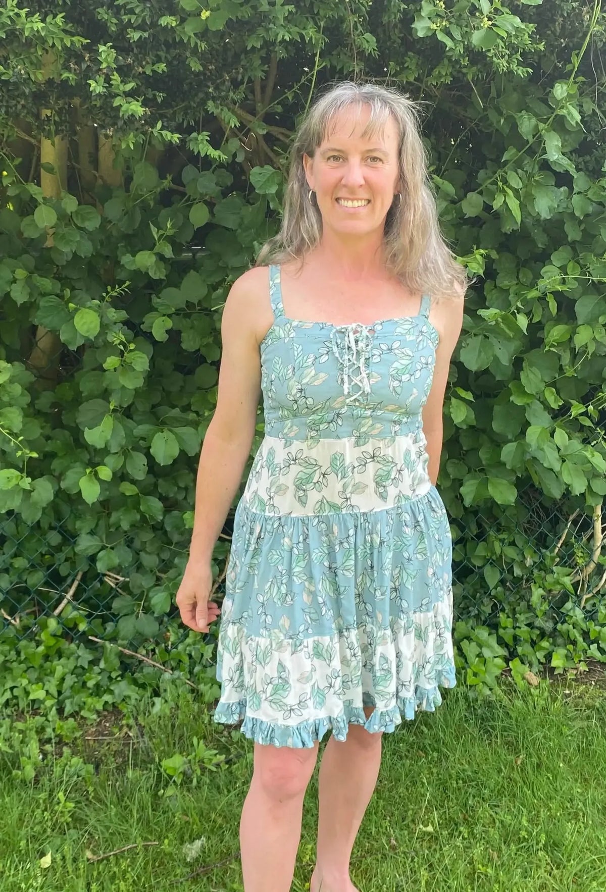 A woman in a light blue and white floral dress stands in front of green foliage.