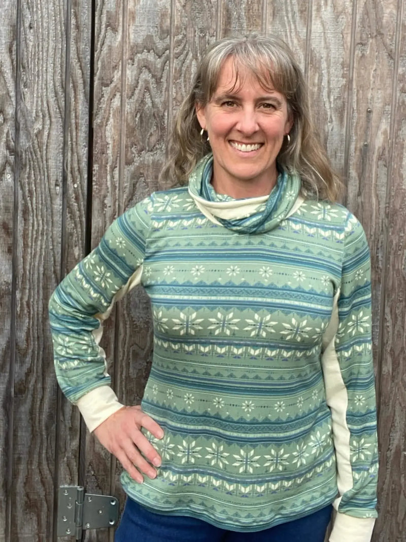 A woman smiles, wearing a green and blue patterned long-sleeve top with a cowl neck.