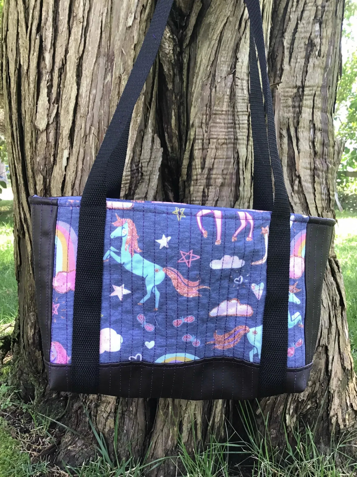A navy unicorn print tote bag with black straps hangs against a tree trunk.