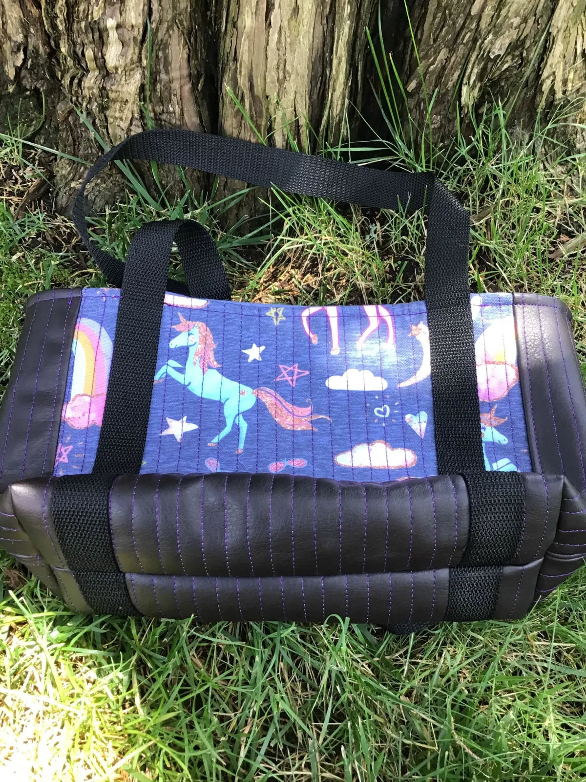 A navy unicorn print bag with black straps and purple stitching sits on green grass.